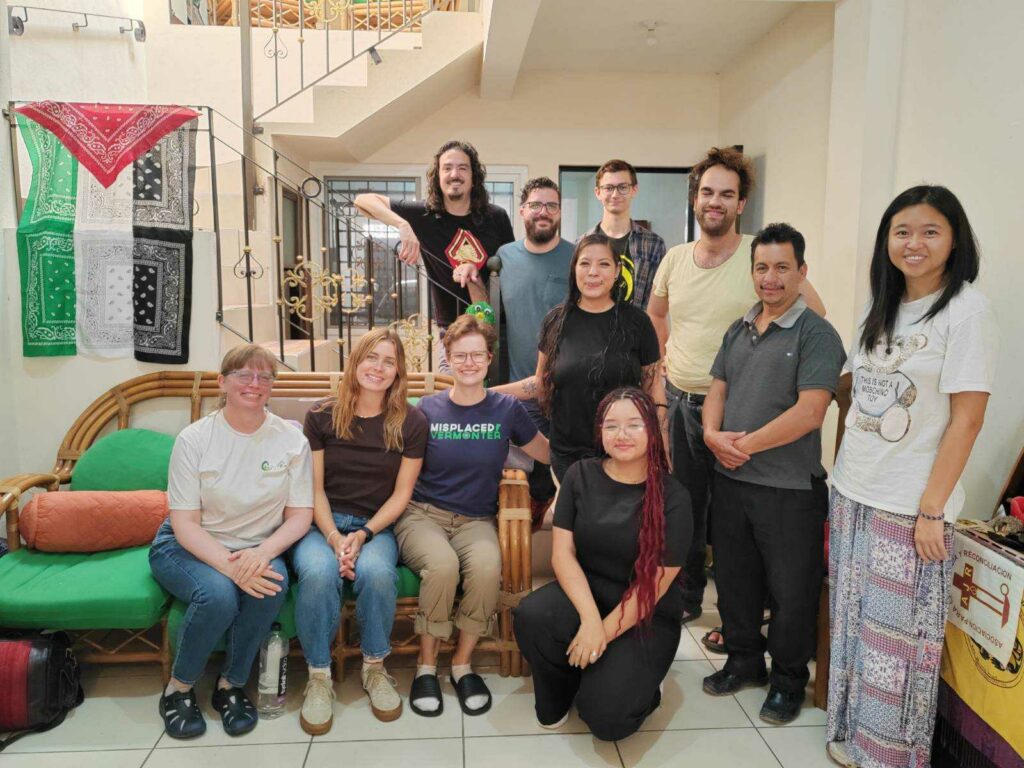 Eleven people pose together. Three of them sit on a green couch, one kneels, and the rest stand by a stairway. On the left hangs a Palestinian flag and on the right a banner with the letters AJR on a cross. Once personas posan juntas. Tres de ellas están sentadas en un sofá verde, una arrodillada y el resto de pie junto a una escalera. A la izquierda cuelga una bandera palestina y a la derecha una pancarta con las letras AJR sobre una cruz.