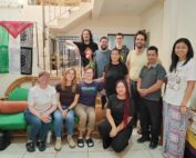 Eleven people pose together. Three of them sit on a green couch, one kneels, and the rest stand by a stairway. On the left hangs a Palestinian flag and on the right a banner with the letters AJR on a cross. Once personas posan juntas. Tres de ellas están sentadas en un sofá verde, una arrodillada y el resto de pie junto a una escalera. A la izquierda cuelga una bandera palestina y a la derecha una pancarta con las letras AJR sobre una cruz.