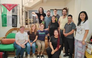 Eleven people pose together. Three of them sit on a green couch, one kneels, and the rest stand by a stairway. On the left hangs a Palestinian flag and on the right a banner with the letters AJR on a cross. Once personas posan juntas. Tres de ellas están sentadas en un sofá verde, una arrodillada y el resto de pie junto a una escalera. A la izquierda cuelga una bandera palestina y a la derecha una pancarta con las letras AJR sobre una cruz.