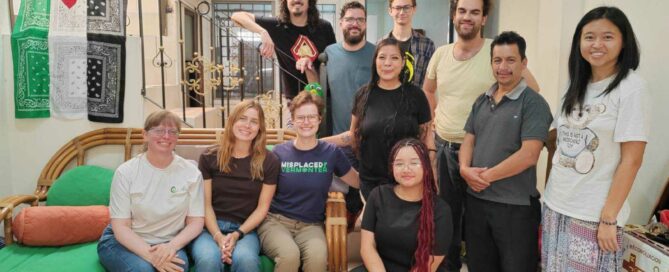 Eleven people pose together. Three of them sit on a green couch, one kneels, and the rest stand by a stairway. On the left hangs a Palestinian flag and on the right a banner with the letters AJR on a cross. Once personas posan juntas. Tres de ellas están sentadas en un sofá verde, una arrodillada y el resto de pie junto a una escalera. A la izquierda cuelga una bandera palestina y a la derecha una pancarta con las letras AJR sobre una cruz.