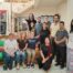 Eleven people pose together. Three of them sit on a green couch, one kneels, and the rest stand by a stairway. On the left hangs a Palestinian flag and on the right a banner with the letters AJR on a cross. Once personas posan juntas. Tres de ellas están sentadas en un sofá verde, una arrodillada y el resto de pie junto a una escalera. A la izquierda cuelga una bandera palestina y a la derecha una pancarta con las letras AJR sobre una cruz.