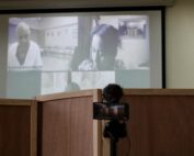 In the foreground you can see a camera and a microphone, behind the screens to protect the identity of the witnesses and in the background you can see the online presence of the accused Lucas Garcia on a screen on the left, wearing a white coat and on the right you can see a woman