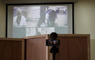 In the foreground you can see a camera and a microphone, behind the screens to protect the identity of the witnesses and in the background you can see the online presence of the accused Lucas Garcia on a screen on the left, wearing a white coat and on the right you can see a woman