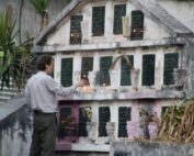 A man lights a candle in front of a concrete mausoleum with two dozen black marble inscriptions including a bible verse and praying hands. Lit candles and flowers line the graves.