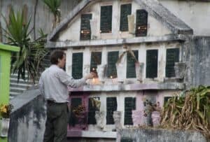 A man lights a candle in front of a concrete mausoleum with two dozen black marble inscriptions including a bible verse and praying hands. Lit candles and flowers line the graves.