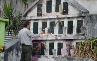 A man lights a candle in front of a concrete mausoleum with two dozen black marble inscriptions including a bible verse and praying hands. Lit candles and flowers line the graves.
