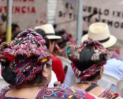 en el fondo se ven unas mantas, en primer plano los tocados de dos mujeres ixiles sentadas en el publico. // At a second plane there are some banners and at the first plane, the traditional hair style of two ixil women