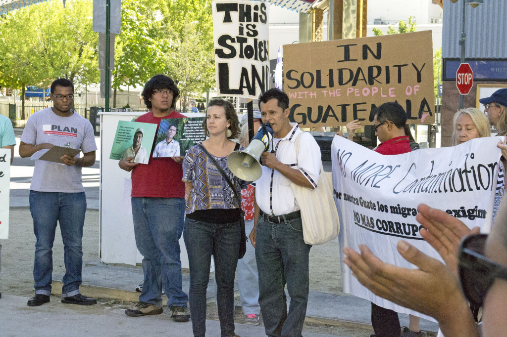 NISGUA tour speaker Llan Carlos speaks into a megaphone at a protest in Reno, Nevada. Behind him are people holding signs, "This is stolen land," and "In solidarity with the people of Guatemala."