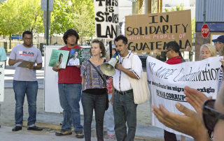 NISGUA tour speaker Llan Carlos speaks into a megaphone at a protest in Reno, Nevada. Behind him are people holding signs, "This is stolen land," and "In solidarity with the people of Guatemala."