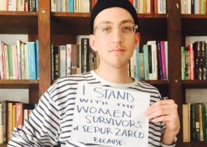 A person stand in front of a bookshelf with a sign that read "I stand with the women survivors of Sepur Zarco."