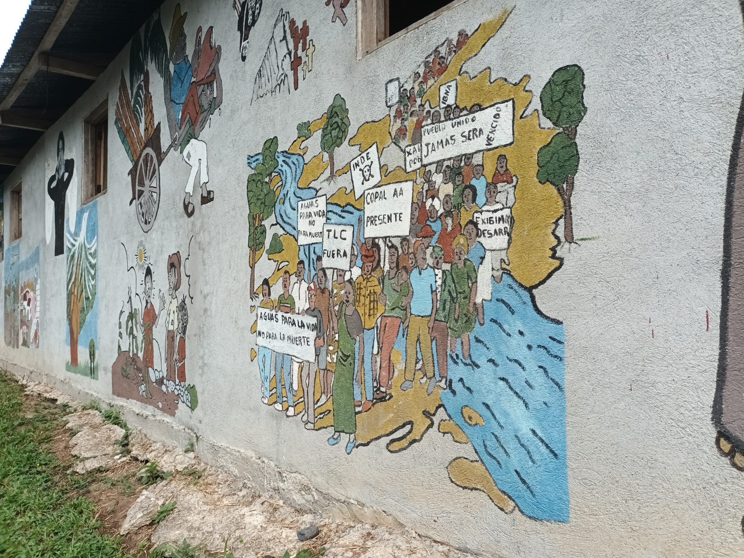 Mural painted on the wall of a building, depicting scenes of rural life and social struggles in Guatemala. On the right, a group of people marches holding signs with messages like “Water for life, not for death,” “No to the free trade agreement,” and “The people united will never be defeated,” over a map of the country. On the left, agricultural workers, religious figures, and natural elements like trees and corn are depicted.