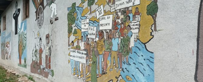 Alt Text: La pared del Instituto Basic es gris. El mural principal muestra a un grupo de personas cruzando un río azul, vestidas de verde, amarillo, naranja y blanco. Hay árboles alrededor del río y el suelo sobre el que están las personas del mural es amarillo.