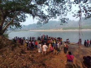 Alt Text: A far off photo of a group of around 60 people gathered at the Rio Chixoy. The group is in the foreground with a big tree arching over the group on the left, the river is in the background. The other side of the shore is furthest back, including a sandy bank and many trees. The most visible costume is a painted monarch butterfly sign on someone’s back. Four people are on stilts, one of whom has a pink wig.