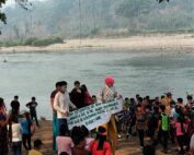 Gran grupo de aproximadamente 50 personas, principalmente niños y jóvenes, congregados en la orilla pedregosa de un río. Varias personas llevan disfraces coloridos, destacando una figura con peluca rosa brillante y camisa de lunares. Un grupo sostiene una pancarta blanca con texto en español. El paisaje muestra un río de aguas tranquilas, una playa de arena al otro lado, y al fondo colinas cubiertas de vegetación tropical envueltas en neblina matutina. Ramas de árboles enmarcan la parte superior de la imagen.