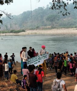 Gran grupo de aproximadamente 50 personas, principalmente niños y jóvenes, congregados en la orilla pedregosa de un río. Varias personas llevan disfraces coloridos, destacando una figura con peluca rosa brillante y camisa de lunares. Un grupo sostiene una pancarta blanca con texto en español. El paisaje muestra un río de aguas tranquilas, una playa de arena al otro lado, y al fondo colinas cubiertas de vegetación tropical envueltas en neblina matutina. Ramas de árboles enmarcan la parte superior de la imagen.