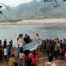 Gran grupo de aproximadamente 50 personas, principalmente niños y jóvenes, congregados en la orilla pedregosa de un río. Varias personas llevan disfraces coloridos, destacando una figura con peluca rosa brillante y camisa de lunares. Un grupo sostiene una pancarta blanca con texto en español. El paisaje muestra un río de aguas tranquilas, una playa de arena al otro lado, y al fondo colinas cubiertas de vegetación tropical envueltas en neblina matutina. Ramas de árboles enmarcan la parte superior de la imagen.