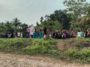 Grupo numeroso de personas marchando al aire libre en un entorno rural, algunas con trajes coloridos, zancos y tocados de plumas, rodeadas de vegetación y árboles tropicales.