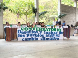 Grupo de personas sentadas detrás de mesa con textiles coloridos, sosteniendo pancarta blanca que dice "CONVERSATORIO Desafíos y luchas de los pueblos ante el cambio climático". Escena en espacio techado con columnas de concreto y vegetación tropical de fondo.