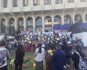 In the foreground, a group of people seen from behind, some holding tripods and cameras. At the center, a ceremonial altar with lit candles and flowers arranged in a circle on pine needles. In the background, a large group gathers on the steps—many women wear traditional clothing and hold posters and papers. Behind them, a building stands behind a metal fence.