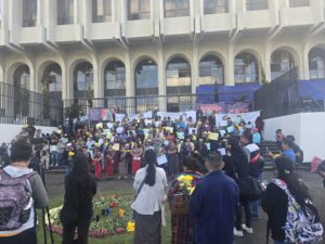 In the foreground, a group of people seen from behind, some holding tripods and cameras. At the center, a ceremonial altar with lit candles and flowers arranged in a circle on pine needles. In the background, a large group gathers on the steps—many women wear traditional clothing and hold posters and papers. Behind them, a building stands behind a metal fence.