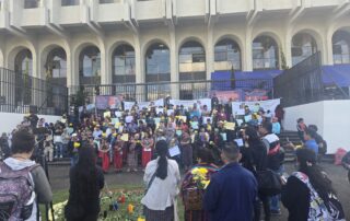 In the foreground, a group of people seen from behind, some holding tripods and cameras. At the center, a ceremonial altar with lit candles and flowers arranged in a circle on pine needles. In the background, a large group gathers on the steps—many women wear traditional clothing and hold posters and papers. Behind them, a building stands behind a metal fence.