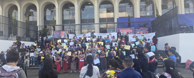 In the foreground, a group of people seen from behind, some holding tripods and cameras. At the center, a ceremonial altar with lit candles and flowers arranged in a circle on pine needles. In the background, a large group gathers on the steps—many women wear traditional clothing and hold posters and papers. Behind them, a building stands behind a metal fence.