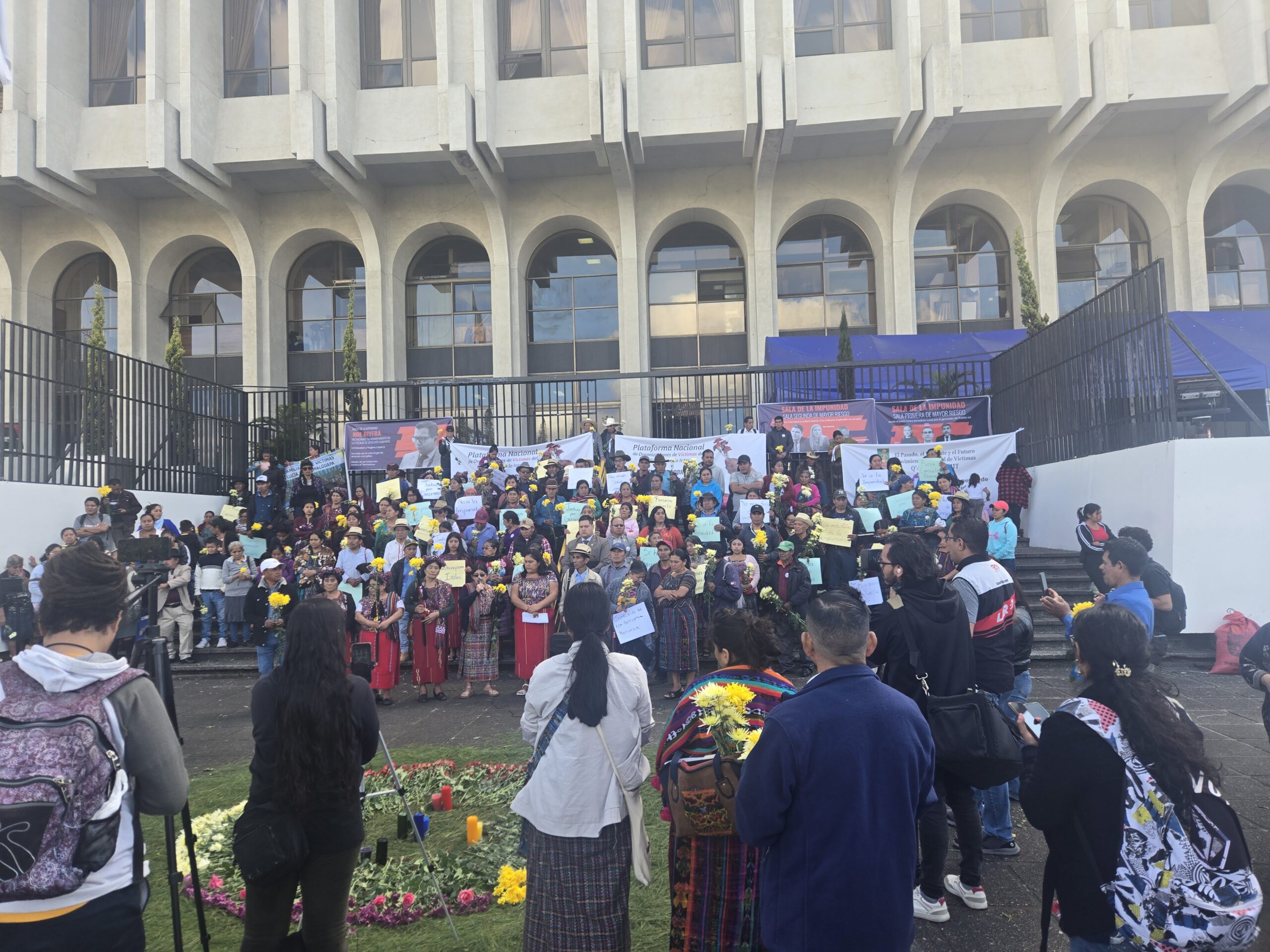 In the foreground, a group of people seen from behind, some holding tripods and cameras. At the center, a ceremonial altar with lit candles and flowers arranged in a circle on pine needles. In the background, a large group gathers on the steps—many women wear traditional clothing and hold posters and papers. Behind them, a building stands behind a metal fence.