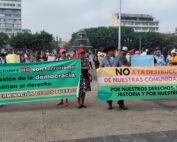 Two green and yellow banners with black, red, and white letters are visible. A group of people wearing caps and hats are carrying them. In the background, there is a fountain and buildings.