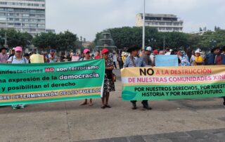 Two green and yellow banners with black, red, and white letters are visible. A group of people wearing caps and hats are carrying them. In the background, there is a fountain and buildings.