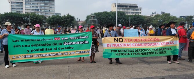 Two green and yellow banners with black, red, and white letters are visible. A group of people wearing caps and hats are carrying them. In the background, there is a fountain and buildings.