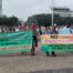 Two green and yellow banners with black, red, and white letters are visible. A group of people wearing caps and hats are carrying them. In the background, there is a fountain and buildings.