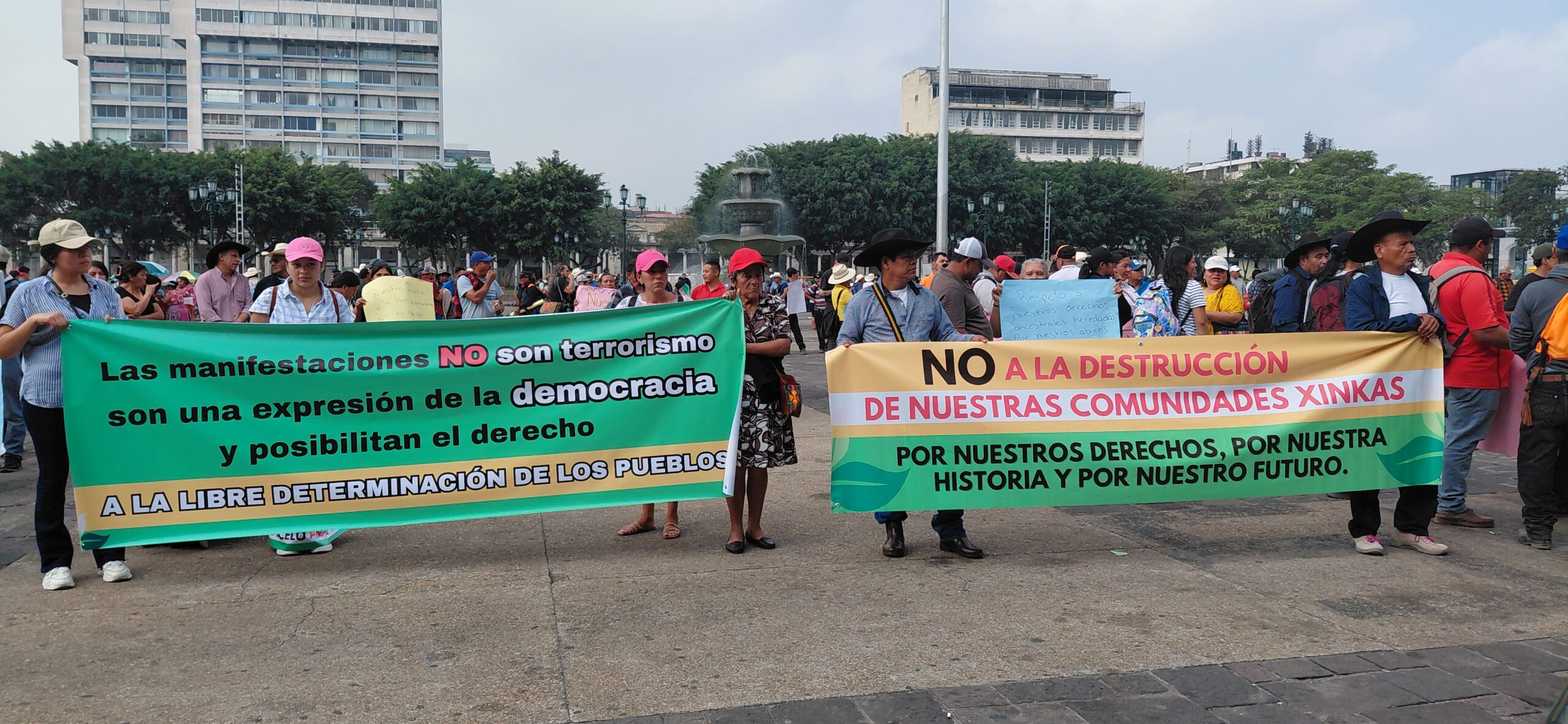 Two green and yellow banners with black, red, and white letters are visible. A group of people wearing caps and hats are carrying them. In the background, there is a fountain and buildings.