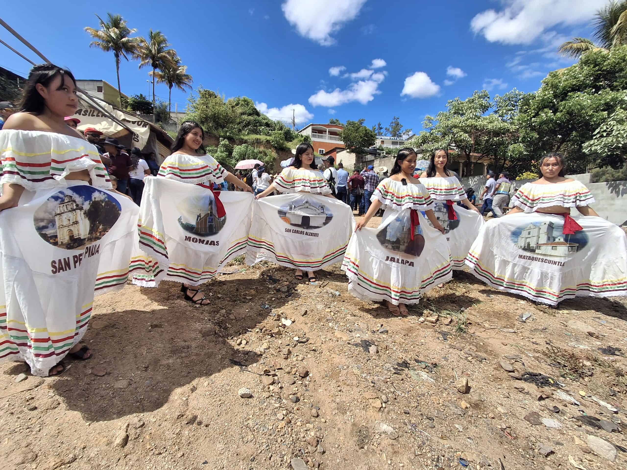 A group of young women dressed in traditional white dresses with colorful stripes, each featuring a printed image and name of a municipality from Jalapa, Guatemala (e.g., San Pedro Pinula, Monjas, San Carlos Alzatate, Jalapa, Mataquescuintla). They are standing in a semi-circle outdoors during a cultural event, with a crowd and palm trees in the background under a bright blue sky.