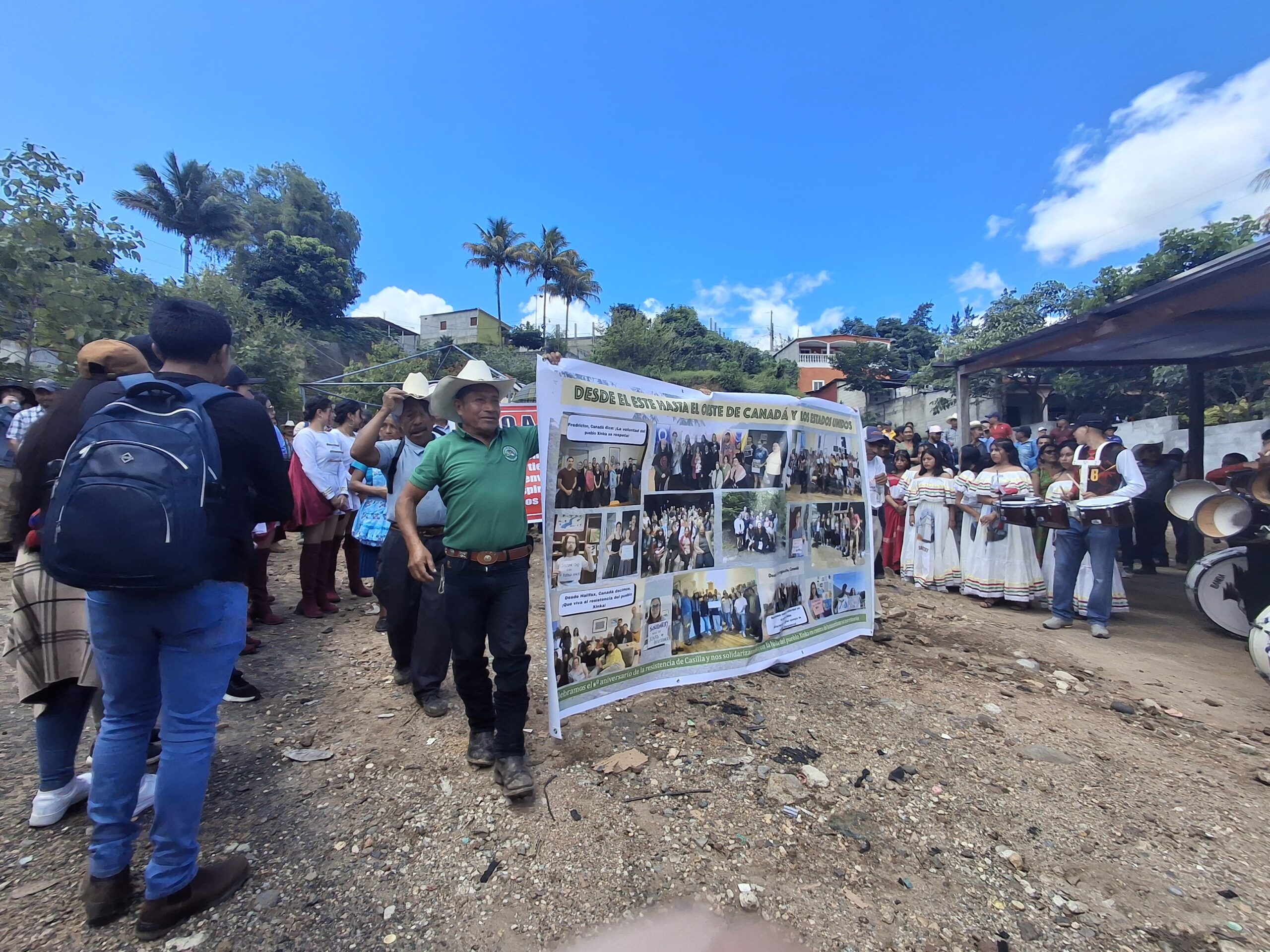 A group of people, including men in traditional hats and young women in traditional white dresses with colorful stripes, stand around a large display board with photos and text about international solidarity, particularly from Canada and the U.S. The event takes place outdoors under a bright blue sky, with palm trees, houses, and a covered structure visible in the background. A marching band stands nearby with drums.