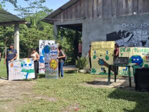 Varias personas jóvenes sostienen carteles pintados a mano con mensajes y dibujos sobre la protección del agua y el cuidado del planeta, de pie frente a un edificio de madera y concreto, con un mural en la pared y vegetación alrededor.