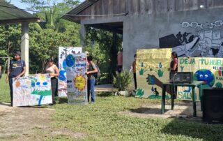 Varias personas jóvenes sostienen carteles pintados a mano con mensajes y dibujos sobre la protección del agua y el cuidado del planeta, de pie frente a un edificio de madera y concreto, con un mural en la pared y vegetación alrededor.