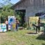 Varias personas jóvenes sostienen carteles pintados a mano con mensajes y dibujos sobre la protección del agua y el cuidado del planeta, de pie frente a un edificio de madera y concreto, con un mural en la pared y vegetación alrededor.