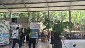 Evento bajo un techo de lámina corrugada donde varias personas sostienen carteles y pancartas, algunos con símbolos de calaveras y textos que incluyen palabras como "MATA" y referencias ambientales. Hay personas de diferentes edades presentes, algunas de pie con los carteles, otras sentadas observando. El espacio tiene piso de concreto y está rodeado de abundante vegetación tropical incluyendo plantas de plátano. Se observa un perro caminando por el área y equipos como altavoces en el espacio cubierto.