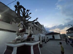 Sunset over a concrete courtyard, foregrounded by a statue depicting a scene from the Popol Vuy, including two seated figures and a tree of many fruits. Children play football in the background.