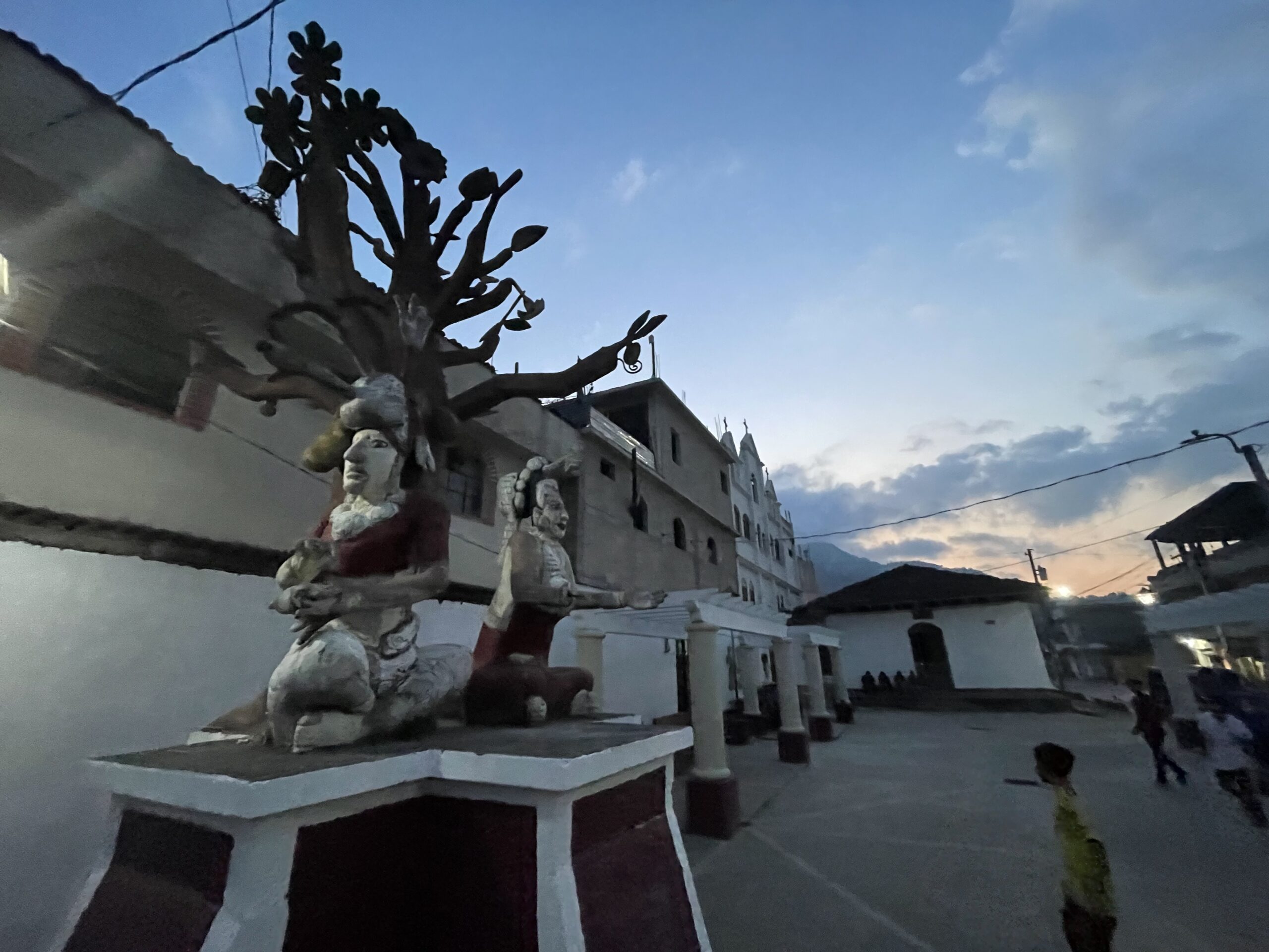 Atardecer sobre un patio de concrete, en primer plano por una estatua que representa una escena del Popul Vuh, incluyendo dos figuras sentadas y un árbol de varias frutas. Los niños juegan al fútbol de fondo.