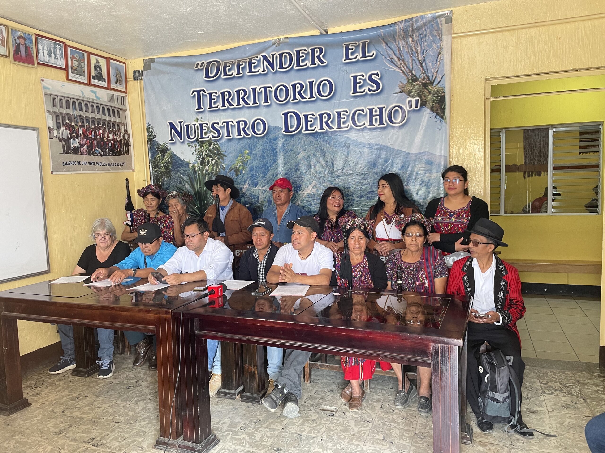 A dozen seated and standing figures hold a press conference. People are dressed in a mix of casual clothing and tradition Ixil attire. Behind the figures is a banner depicting a mountain range, reading "Defender el Territorio es Nuestro Derecho" (Defending [our] territory is our right)