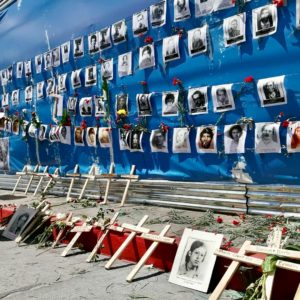 Picture of a giant poster with Guatemalan national colours, covered by pictures of the dissapeared and murder during the Internal Armed Conflict along with red carnationsand crosses with their names.