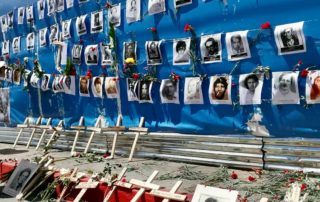Picture of a giant poster with Guatemalan national colours, covered by pictures of the dissapeared and murder during the Internal Armed Conflict along with red carnationsand crosses with their names.