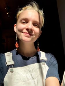 Oli a white non binary presenting person, smiling for the camera, wearing a beige overall, blue navy shirt and orange earnings.
