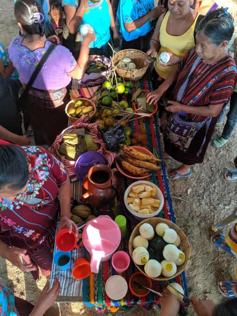 Women, many in traditional Maya Mam clothing, gather around a brightly colored table heaping with fresh fruit and drinks. 