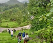 In the right foreground we can see some trees, then a road and several people walking downhill. In the background you can see another house and a very green landscape. // Se observa en primer plano del lado derecho unos árboles, después un camino y varias personas bajando. En el fondo se ven otra casa y un paisaje muy verde.
