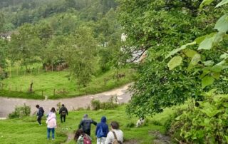 In the right foreground we can see some trees, then a road and several people walking downhill. In the background you can see another house and a very green landscape. // Se observa en primer plano del lado derecho unos árboles, después un camino y varias personas bajando. En el fondo se ven otra casa y un paisaje muy verde.