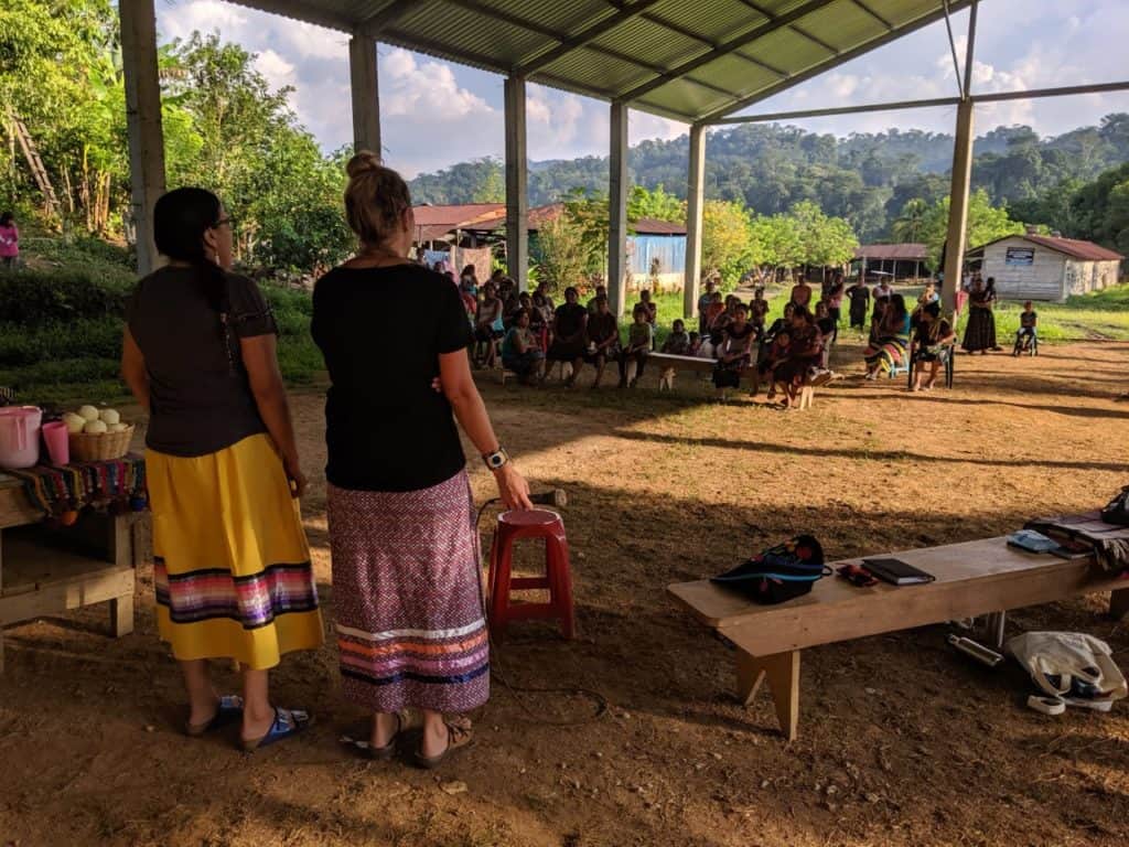 Two women in ribbon skirts stand before 40 people sitting in chairs. Their backs are to the camera and the sun is setting.