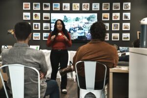 Una mujer está de pie al frente de un salón dando una presentación a una pequeña audiencia sentada. Lleva una camisa roja de manga larga y habla con las manos ligeramente levantadas. Detrás de ella, una pantalla grande muestra una escena al aire libre con personas y pancartas. La pared alrededor de la pantalla está decorada con una cuadrícula de fotos enmarcadas. Los miembros de la audiencia están sentados en sillas metálicas blancas, vistos desde atrás.