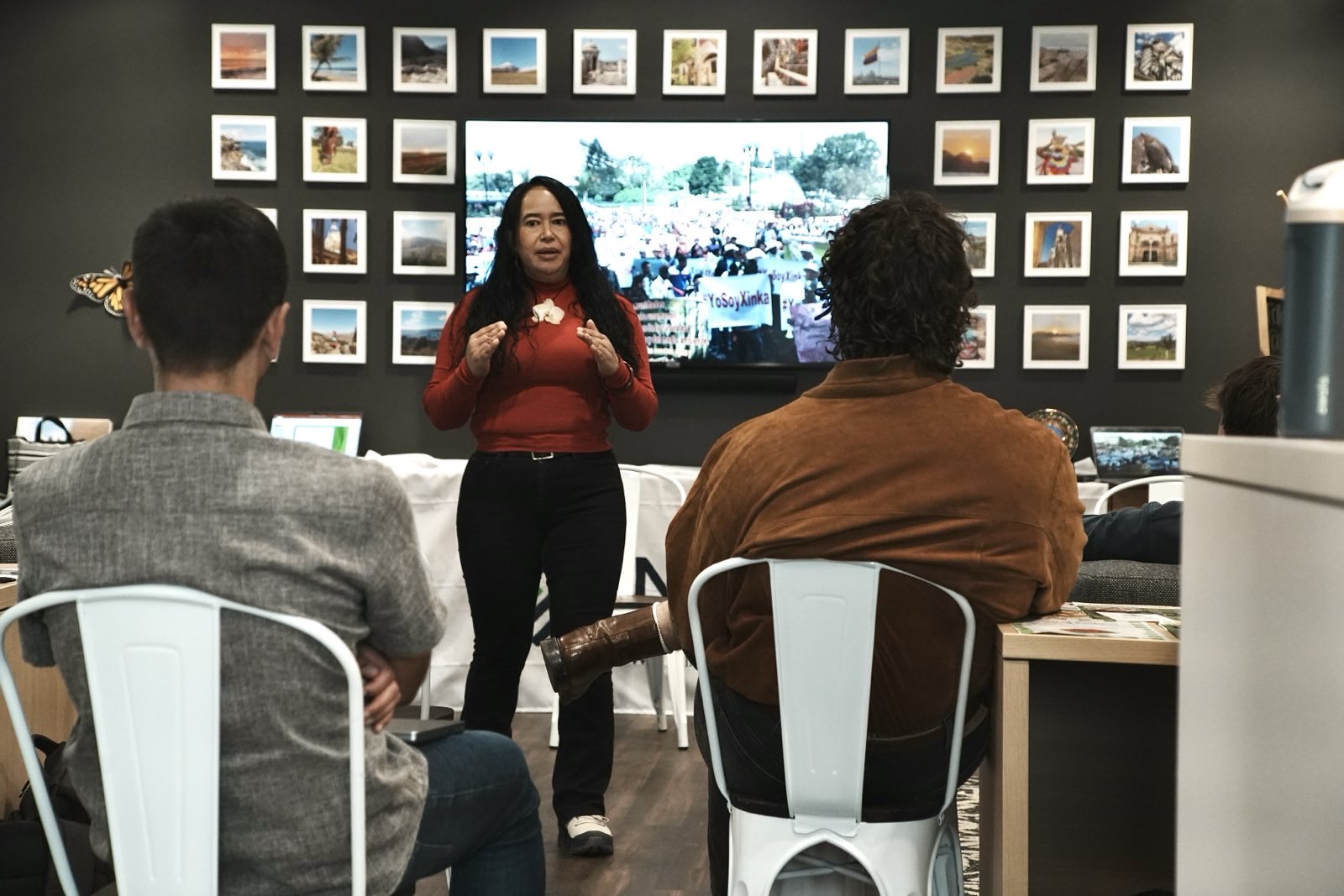A woman stands at the front of a room giving a presentation to a small seated audience. She wears a red long-sleeve shirt and speaks with her hands slightly raised. Behind her, a large screen displays an outdoor scene with people and banners. The wall around the screen is decorated with a grid of framed photos. The audience members sit on white metal chairs, seen from behind.